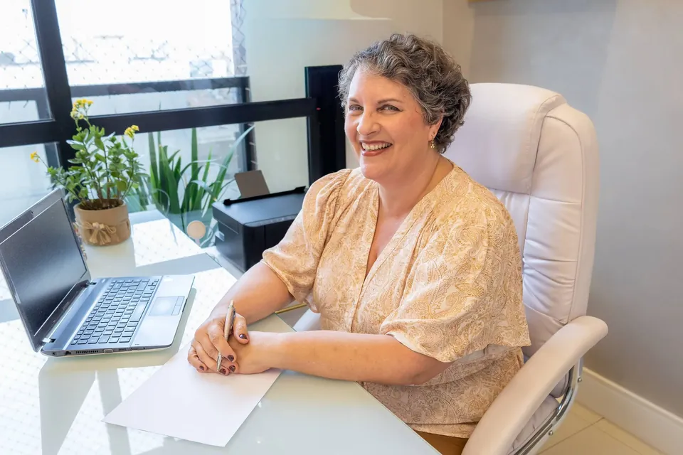 Dra. Patrícia sentada à mesa de atendimento, sorrindo, com plantas e uma janela de luz natural ao fundo.